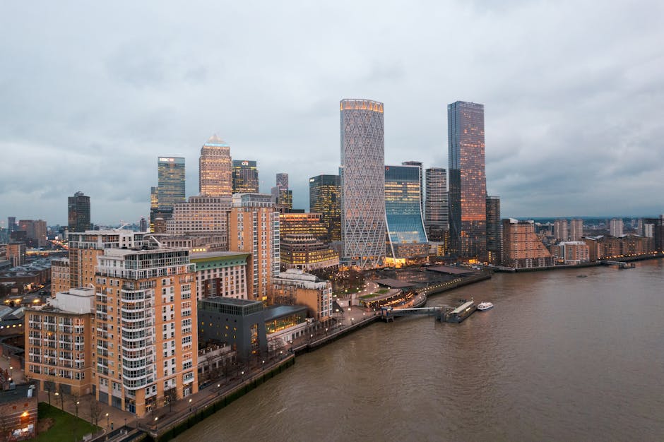 A wide view of London's Canary Wharf skyline at dusk, featuring modern high-rise office buildings with glass facades reflecting the overcast sky. The prominent buildings include a topped-off tower wit