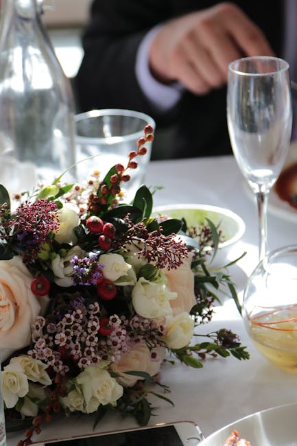 A close-up of a floral arrangement featuring a mix of white roses, purple lilacs, sprigs of red berries, and various greenery, arranged in a loose, natural style on a white tablecloth. The bouquet is 