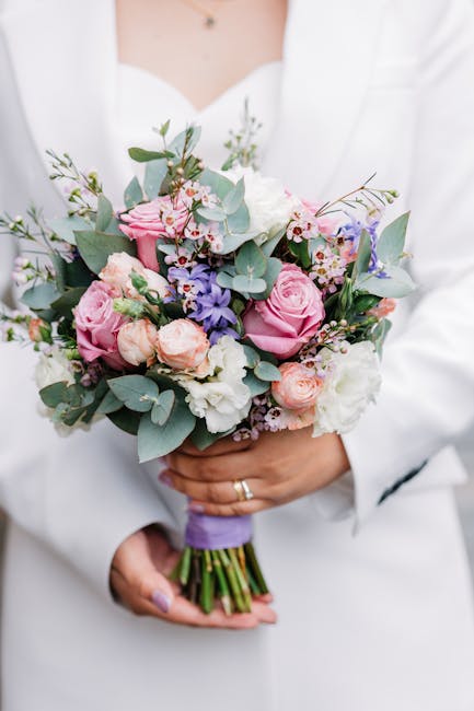 A person dressed in a white outfit is holding a floral bouquet featuring pink roses, white lisianthus, light purple delphiniums, and sprigs of eucalyptus. The bouquet is wrapped with purple ribbon aro