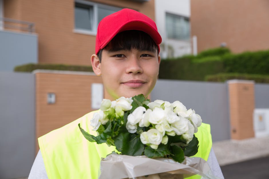 A young male delivery person wearing a red cap and a high-visibility yellow vest is standing outdoors in front of modern residential buildings with brown brick walls and greenery. He is holding a bouq