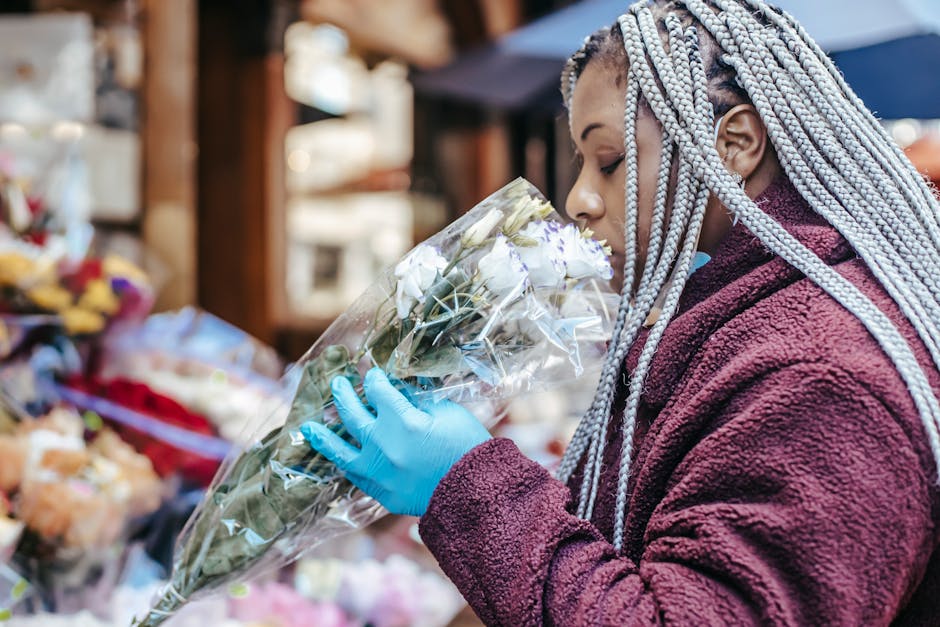 A person with braided, silver-gray hair and wearing a maroon fleece jacket is holding a bouquet of white roses wrapped in clear cellophane. They are wearing gloves and appear to be inspecting or smell